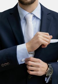 Laivastonsininen puku, vaaleansininen paita, valkoinen timanttikuviollinen solmio, hopeiset kalvosinnapit ja musta kello, jossa on raidallinen hihna.