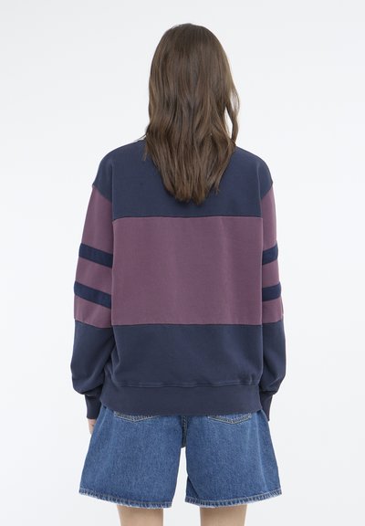 Hanorac din bumbac cu dungi orizontale bleumarin și burgund, având mâneci lungi și un croi relaxat, asortat cu șorturi din denim.