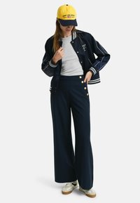 Veste de bombardier bleu marine avec des rayures blanches, t-shirt gris, pantalon bleu marine taille haute avec des boutons dorés, et casquette jaune avec des accents noirs.