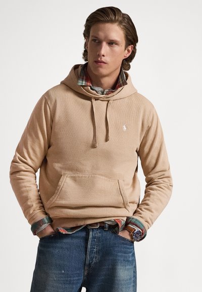 Sudadera con capucha de color beige con un bolsillo frontal tipo canguro, cordón ajustable y logotipo bordado. Combina con una camisa de cuadros y se lleva con jeans azules.