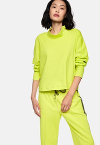 Frau mit schwarzen Haaren, die ein leuchtend gelbes langärmliges Oberteil und passende gestreifte Pants mit einem Kordelzug und seitlichen Streifen trägt.