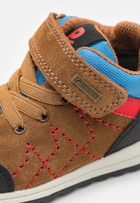 Pantof high-top din suede maro, cu plasă albastră și accente roșii, având o curea cu arici, cusături decorative și o talpă din cauciuc negru.