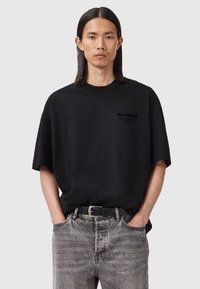 Schwarzes oversized T-Shirt mit dezenter "ALLSAINTS"-Markenkennzeichnung, kombiniert mit grauen Denim-Jeans und einem schwarzen Gürtel. Glattes Material und lockere Passform.