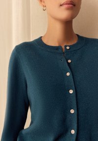 Modro-zeleni cardigan iz mehke tkanine z okroglim izrezom, ki ima pet gumbov spredaj in nežen teksturni vzorec.