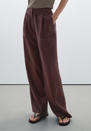 Personne portant un pantalon marron ample et des tongs noires, debout avec une main dans la poche, sur un fond uni.