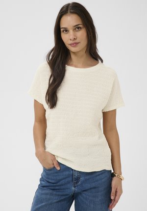 Jeune femme aux cheveux longs et foncés portant un haut texturé à manches courtes de couleur écru, un jean bleu et une montre-bracelet noire et dorée.