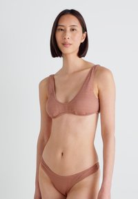 Hnedý textúrovaný bikini set, ktorý obsahuje podprsenku s hlbokým výstrihom a zodpovedajúce nízko strihané nohavičky. Tkanina vyzerá hladko a má hrubé ramienka pre lepšiu oporu.