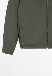 Giacca verde oliva con cerniera frontale, orlo elastico e tasca laterale sinistra. Manica e metà inferiore della giacca visibili.