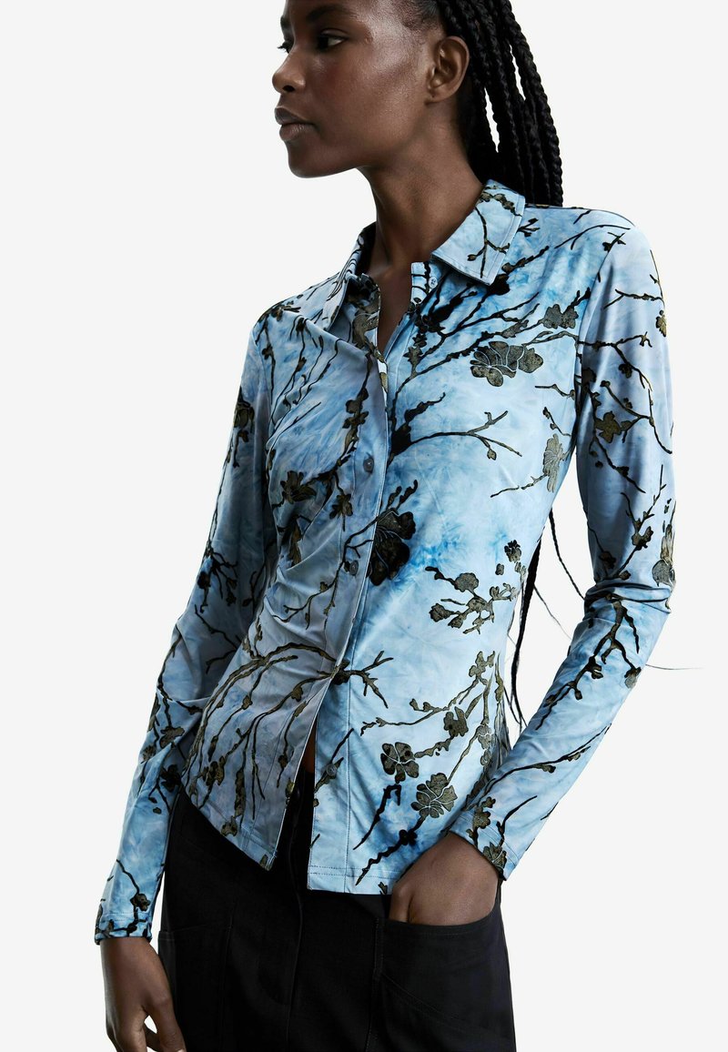 Nainen, jolla on lettiin punottu tukka, pukeutunut istuvaan, pitkähihainen sininen paita, jossa mustat kukka- ja oksakuvioinnit, yhdistetty mustiin housuihin, käsi taskussa.