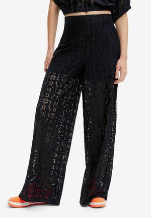 Vrouw draagt hooggetailleerde zwarte brede broek met bloemenkant, cropped zwart topje en feloranje sneakers, staand tegen een witte achtergrond.