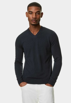 Granatowy sweter w serek wykonany z miękkiego materiału, z długimi rękawami i dopasowanym krojem. Nosi się go na białych spodniach, z rękami w kieszeniach.