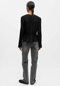 Zwarte geplooide blouse met lange mouwen, een aansluitende taille en een uitlopende zoom, gecombineerd met grijze rechtlopende jeans en zwarte platte schoenen.