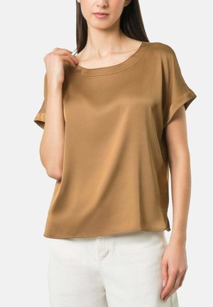Vrouw draagt een losse, kortarmige camelbruine blouse met een ronde halslijn en een witte broek, staand tegen een effen achtergrond.