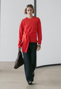 Röd tröja med knytband i midjan, kombinerad med vida mörka jeans. Modellen håller en brun axelväska. Slät textur, avslappnad design.