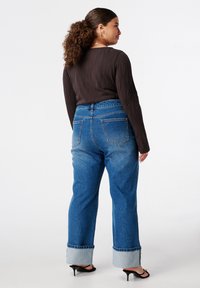 Chemise marron à manches longues côtelées associée à un jean en denim bleu taille haute avec des revers au niveau des poignets et deux poches arrière.