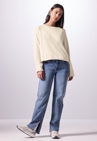 Maglione corto color crema con maniche lunghe, abbinato a jeans blu chiaro a vita alta con spacchi laterali e sneakers bianche.