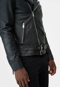 Zwarte leren motorjack met zilveren ritsen, een ceintuur in de taille en een gestructureerde afwerking. Voorzien van ritszakken op de borst en zijkanten.