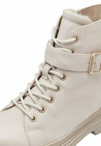 Bottine en simili cuir blanc avec un bout rond, un laçage à l'avant, des œillets dorés et un détail de sangle à boucle au niveau de la cheville. Surface texturée.