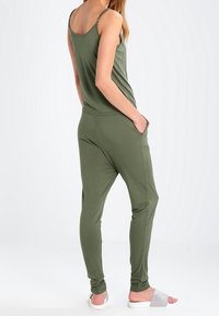 Combinaison verte olive avec des bretelles fines, des jambes fuselées et des poches latérales. Texture lisse et tissu léger ; offre une coupe décontractée.