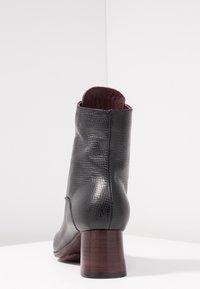 Černá texturovaná kožená kotníková bota se zúženým dřevěným blokem na patě, z pohledu zezadu na bílé ploše a světle šedém pozadí.