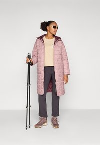 Manteau long rembourré en rose clair avec un motif en losanges, associé à un haut beige, un pantalon sombre et des bottes de randonnée marron, tenant des bâtons de randonnée.