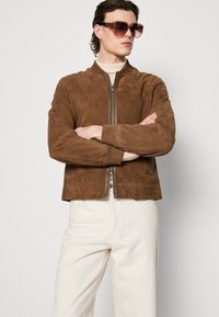 Blouson en daim brun avec fermeture éclair à l'avant, poignets et col côtelés, porté avec un pantalon crème et des lunettes de soleil surdimensionnées.
