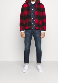 Röd- och svartmönstrad fleecejacka över en jeansskjorta, kombinerad med mörka jeans och vita sneakers. Jackan har en krage och knappar.