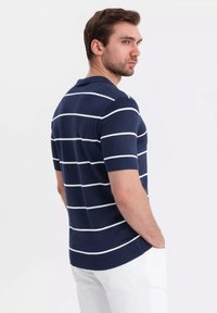 Modrá pruhovaná polo košile s bílými vodorovnými pruhy, krátké rukávy a žebrovaný límeček, spárovaná s bílými kalhotami. Pevná textura látky.