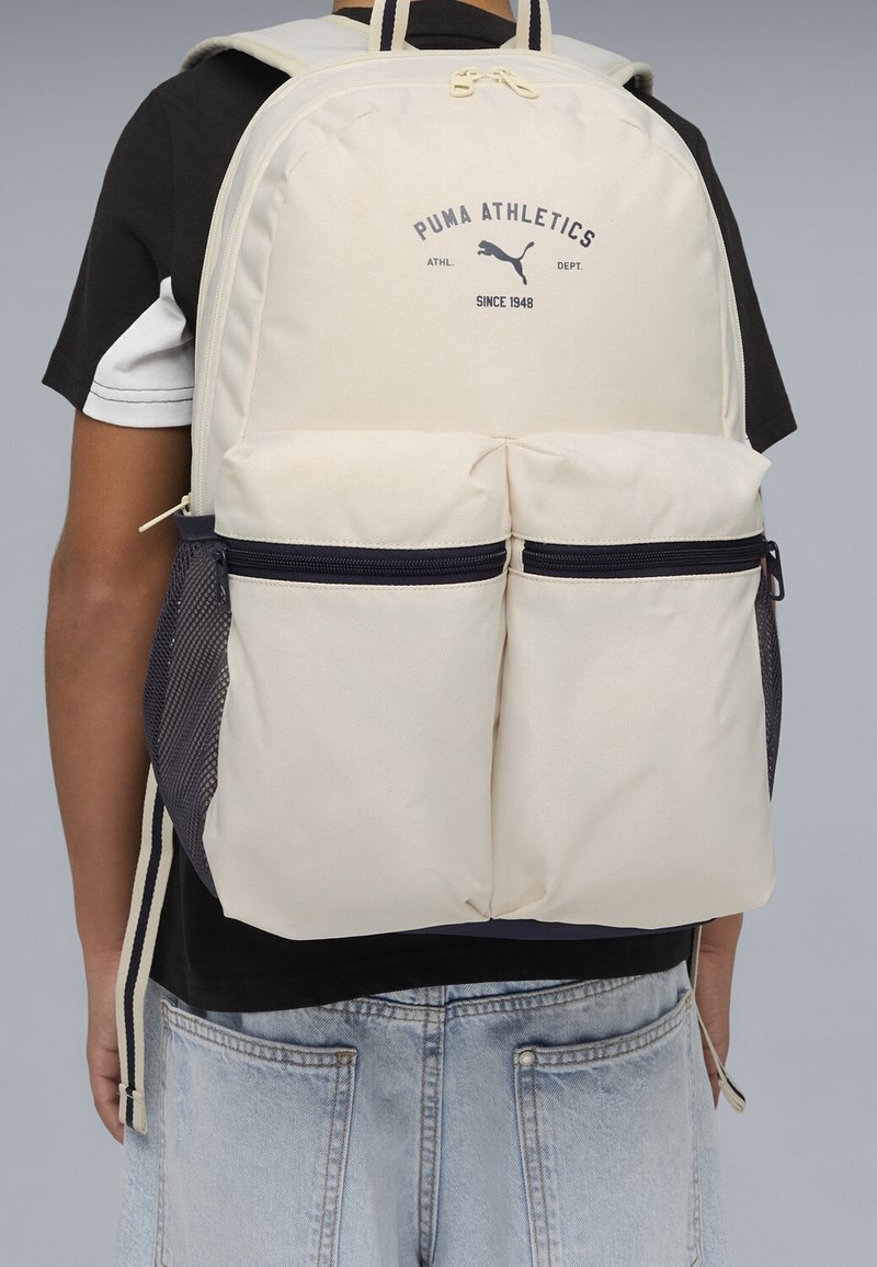 Personne portant un sac à dos beige Puma Athletics avec deux poches avant zippées et des poches latérales en mesh, habillée d'une chemise noire et blanche et d'un jean en denim clair.