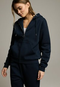 Granatowy rozpinany polar wykonany z miękkiej tkaniny. Posiada kaptur, kieszenie z przodu i sznurki. Połączony z dopasowanymi dresami.