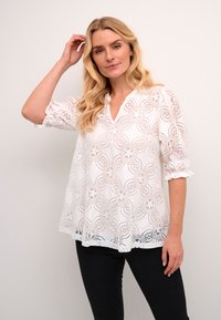 Blouse en dentelle blanche avec un motif floral, des manches volantées et un décolleté en V. Silhouette fluide avec une texture légère.