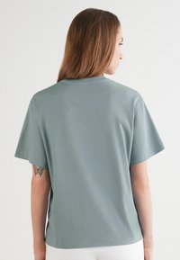 Tričko s krátkým rukávem a kulatým výstřihem z jemné, světle zelené látky. Hladký povrch s uvolněným střihem. Bez viditelných vzorů nebo ozdob.