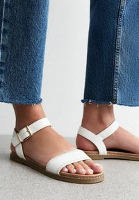 Sandales en cuir blanc avec deux brides, une avec une boucle, semelle plate et détails de couture visibles, portées avec un jean bleu à l'ourlet effiloché.