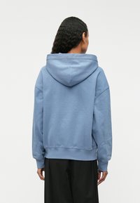 Ανοιχτό μπλε φούτερ με μεγάλη ρυθμιζόμενη κουκούλα, χαλαρούς ώμους και ριμπ μανσέτες. Μαλακή υφή και άνετη εφαρμογή, συνδυασμένο με μαύρο παντελόνι.