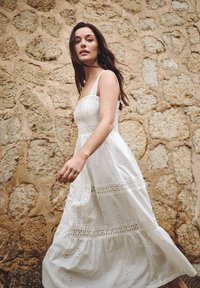 Witte katoenen jurk met een vierkante halslijn, kantdetails en een tiered zoom. De jurk is lichtgewicht en heeft bloemen-uitsnijdingen en verstelbare banden.