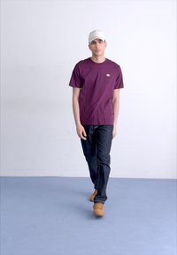 Maroon t-shirt with logo, gray denim jeans, and tan sneakers. The individual is wearing a light gray cap, standing on a blue floor.