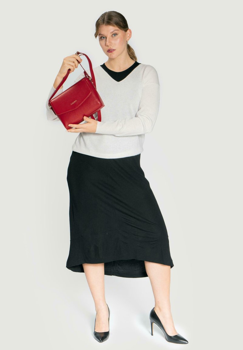 Bolso de mano texturizado en rojo con correa de hombro, sostenido por una persona que lleva un suéter claro y una falda negra, con tacones altos negros y un diseño de escote en V.