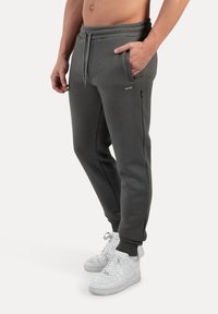 Pantalones de chándal grises con cinturilla elástica, bolsillos laterales y puños acanalados; combinados con zapatillas blancas. Textura suave, diseño casual.