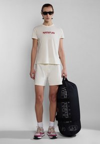 Lichtbeige katoenen T-shirt met rood logo, crèmekleurige shorts en zwarte sporttas. Roze en grijze sneakers, met zwarte zonnebril.
