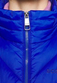 Blue padded jacket featuring a high collar, a central zipper with a metallic pull, and a logo in a reflective accent on the front.
