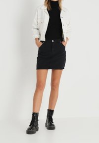 Kvinna i svart polotröja, vit jacka, svart denimkjol och svarta snörade ankellånga stövlar, stående med händerna i fickorna.