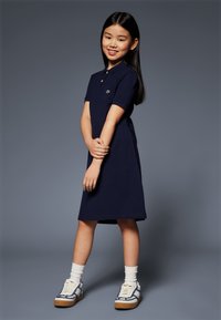 Rochie polo de culoare bleumarin cu mâneci scurte, având un mic emblema pe piept, asortată cu șosete albe și încălțăminte cu accente albastre.