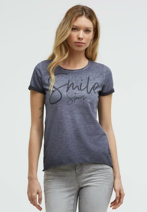 Junge Frau mit langen blonden Haaren, die ein blau-graues T-Shirt mit hochgekrempelten Ärmeln und bedrucktem Text trägt, kombiniert mit hellgrauen Jeans.