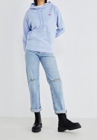 Sweatshirt à capuche bleu clair avec poche avant et cordons, associé à un jean en denim délavé et us é et des bottes militaires noires.