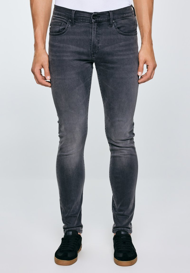 Mann trägt eng anliegende, ausgewaschene schwarze Jeans und schwarze Sneaker mit braunen Sohlen, steht vor einem schlichten hellen Hintergrund.