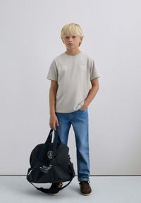 T-shirt en coton gris clair avec des impressions graphiques, jean en denim bleu et un sac de sport noir avec un logo et des détails textuels. Chaussures marron.