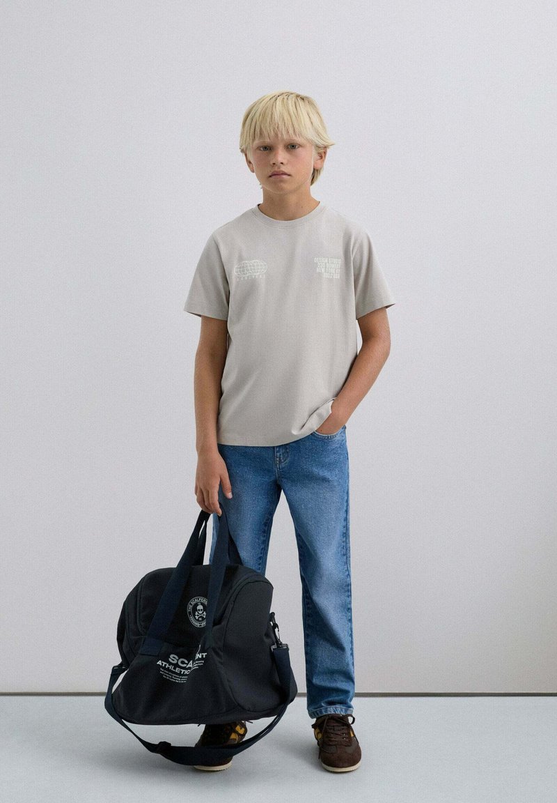 T-shirt en coton gris clair avec des impressions graphiques, jean en denim bleu et un sac de sport noir avec un logo et des détails textuels. Chaussures marron.