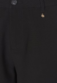 Le tissu noir des pantalons présente une texture lisse, une poche avant et un petit accent en métal doré fixé à la taille.