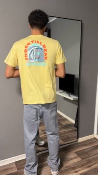 Άνδρας που κοιτάζει σε καθρέφτη τοίχου, φοράει κίτρινη μπλούζα με γραφικό σερφ και γκρι παντελόνια, στέκεται σε ξύλινο πάτωμα σε δωμάτιο με γκρι τοίχους.