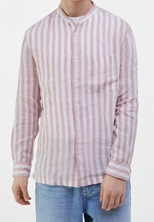 Mann trägt ein rosa-weiß vertikal gestreiftes Hemd mit Stehkragen und blaue Jeans, steht vor weißem Hintergrund.